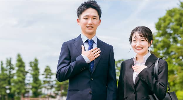 屋外で胸に手を当てて並ぶスーツ姿の男女